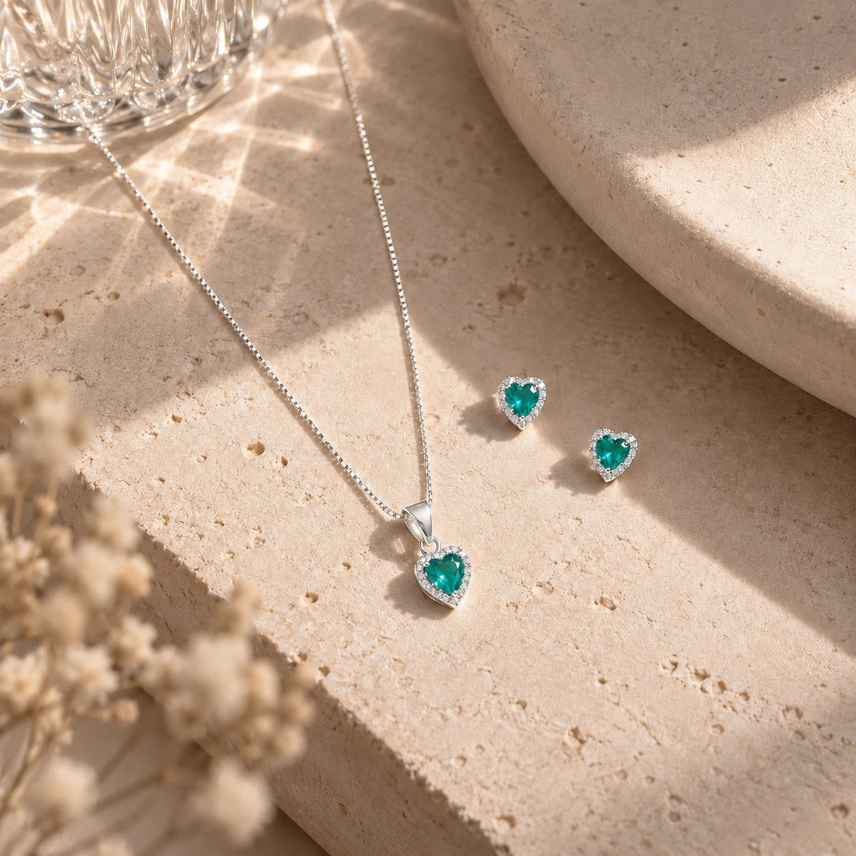 Colar e brincos de coração em prata 925 com zircônia verde esmeralda, sobre pedra bege clara com sombra de folhagem.